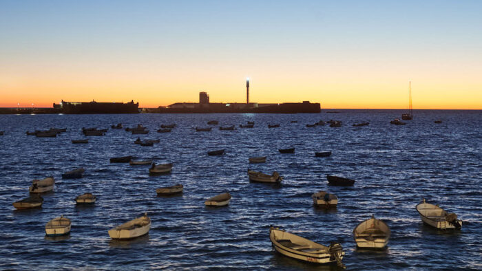 Stranden la Caleta i Cádiz med guppande ekor i förgrunden och Castillo de San Sebastián mot horisonten i solnedgång.