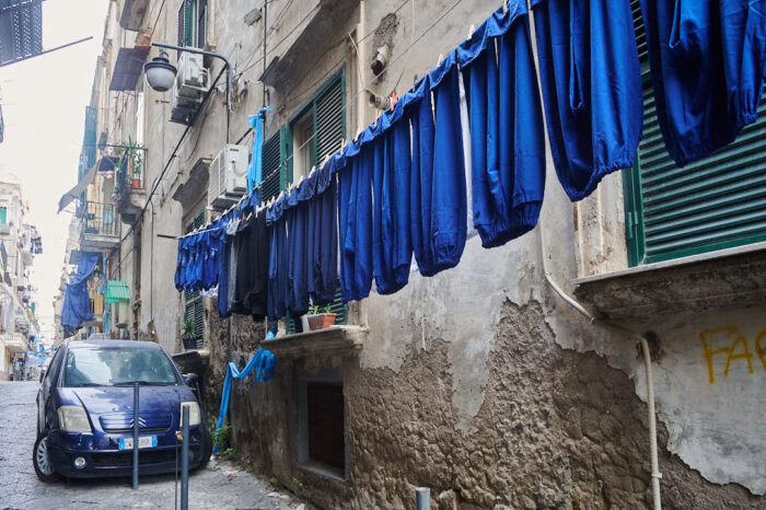 Blå klädplagg på klädstreck och en blå bil på en smal gata i Neapel.